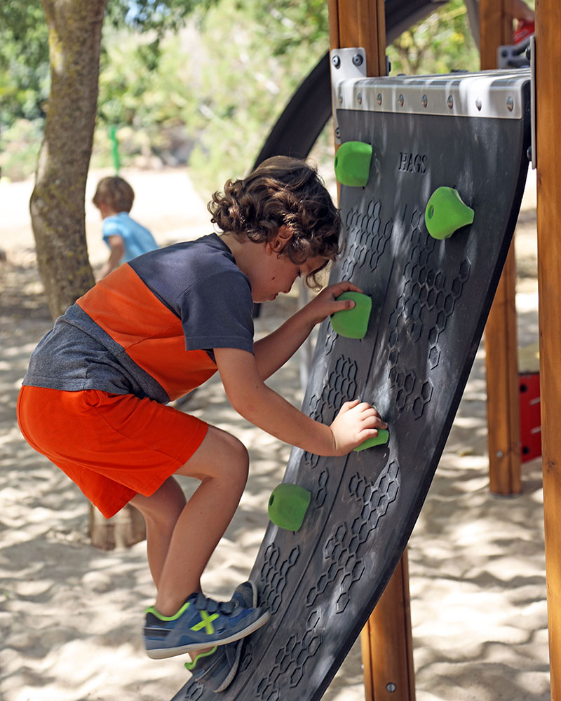 Young boy is using climbing wall with handles to climb up onto a playground unit.